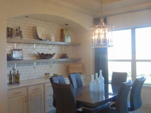 Modern dining area with a white table, gray chairs, and built-in shelving on a tiled wall, illuminated by a chandelier and natural light from a window, featuring craftsmanship by Texas builders.