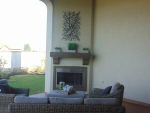 An outdoor covered patio with a fireplace surrounded by wicker chairs, above which hangs an abstract wall art piece and a shelf with green plants, constructed by Texas builders.