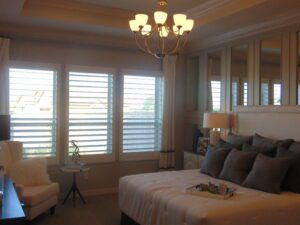 Elegant bedroom with a large bed, white headboard, chandelier, and plantation shutters on windows, meticulously crafted by Texas builders.