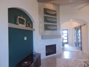 Modern living room with a teal accent wall featuring built-in shelves constructed by Texas builders, a fireplace with a stone surround, and a view into a brightly lit hallway.