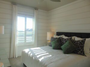 A bright, modern bedroom featuring a large bed with decorative pillows, white shiplap walls crafted by Texas builders, and a window with shutters allowing sunlight to filter in.