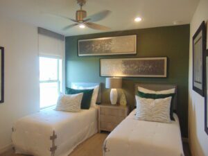 Modern bedroom with two single beds, green accent wall, framed artwork by Texas builders, and a ceiling fan. Brightly lit with natural light from a window.