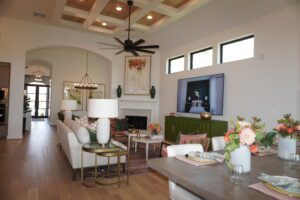 Spacious living room with a beige sofa, white lamps, a green cabinet, and floral accents. Features include a ceiling fan, a fireplace, and wood-beamed ceilings crafted by Texas builders.
