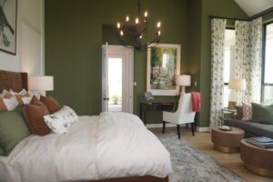 Elegant bedroom featuring a white bed with decorative pillows, green walls, chandelier, and stylish furniture including a desk and chair near a sunlit doorway, designed by Texas builders.