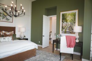 A cozy bedroom featuring a green wall, an open white door, a bed with white linen and brown headboard, two nightstands with lamps, and a framed painting of a tree-lined path by Texas