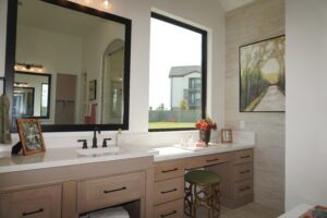 Modern bathroom crafted by Texas builders, featuring a large mirror, dual vanities, and a garden view through a window. A stool and floral decor are visible.