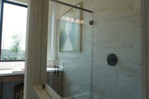 Modern bathroom with glass shower, marble walls, framed artwork by Texas builders, and a view of a window by a desk with a green chair.