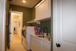 A laundry room with white cabinets, a washing machine, and a sink, featuring a green wall and a hallway view in the background, constructed by Texas builders.