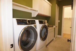 Modern laundry room with green walls, featuring a white front-loading washer and dryer, white cabinetry by Texas builders, and tiled flooring.