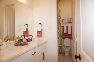 Modern bathroom designed by Texas builders, featuring a white vanity, black faucets, mirror, and a separate toilet area; decorated with red towels and a floral arrangement.