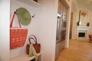 A stylish entryway designed by Texas builders featuring a green hat and an orange-patterned bag hanging on wall hooks, with a brown purse and green scarf on a bench.