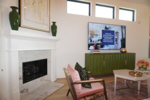 Modern living room with a white marble fireplace, green cabinet, and a flat-screen TV showing a news channel. A stylish chair, sofa, and decorative items completed by Texas builders complete the space.