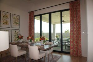 A modern dining room with a sliding glass door leading to a patio, decorated with red curtains, botanical artwork by Texas builders, and a table set for a meal.