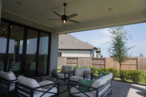 Covered patio area with modern outdoor furniture and ceiling fan, designed by Texas builders, overlooking a backyard with a wooden fence.