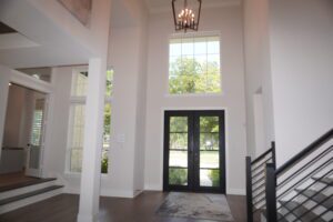 A spacious entryway featuring a high ceiling, white columns, and a double glass door with large windows, leading to a black staircase on the left designed by Texas builders.