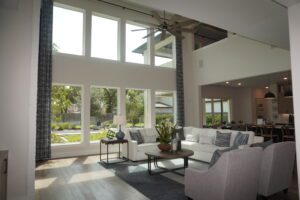 Modern living room with high ceilings, large windows, gray sofas, a ceiling fan, and a view of the adjacent dining area designed by Texas builders.
