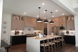 Modern kitchen interior featuring beige cabinetry, stainless steel appliances, a central island with stools, and black pendant lights designed by Texas builders.