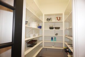 A well-organized walk-in pantry with white shelves built by Texas builders, storing various kitchen items, including bowls, containers, and decorative objects.