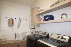 Modern laundry room with white walls, wooden shelves holding towels and jars, and black washing machines, designed by Texas builders.