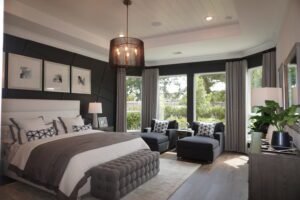 A modern bedroom featuring a large bed with grey bedding, black accent walls, a sitting area with two chairs designed by Texas builders, and a chandelier, leading to a balcony.