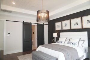 Modern bedroom with a dark panel accent wall, white beddings, a gray throw, and framed photos above the bed. Features barn-style sliding doors and an elegant hanging light fixture designed by Texas builders.