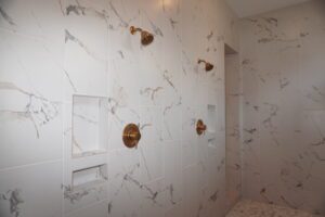 A modern shower with white marble tiles and gold fixtures, including a shower head and knobs, and built-in shelves, crafted by Texas builders.