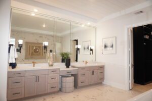 A modern bathroom featuring dual sinks with gray cabinets, large mirrors, and elegant gold faucets, crafted by Texas builders, complemented by white marble walls and floors.