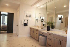 Modern bathroom with double vanity, marble countertops, large mirrors, and white walls. A doorway shows a glimpse of the adjacent bedroom, perfected by Texas builders.