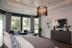 Elegant bedroom with a large bed, dark wooden furniture, and gray armchairs, overlooking a garden through large windows, modern chandelier above, crafted by Texas builders.