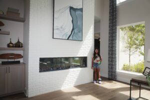 A woman standing in a modern living room with white brick walls, a horizontal fireplace, and a large abstract painting designed by Texas builders.