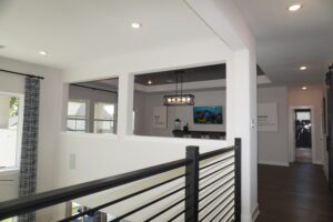 Interior of a modern home crafted by Texas builders, featuring a hallway with dark wood floors, white walls, and a stair railing. A room with a TV is visible through an open window-like wall cut