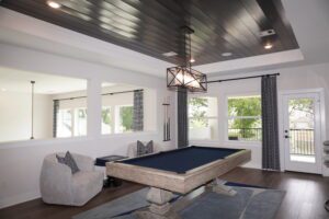 Modern home interior designed by Texas builders, featuring a pool table with blue felt, gray upholstered chairs, dark wood ceiling, and large windows with curtains.