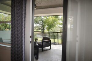 View from an open door showing a small balcony with a single chair, designed by Texas builders, overlooking trees and a clear sky.