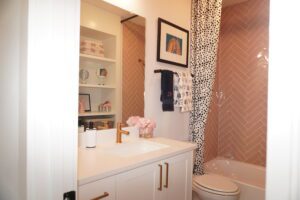 A modern bathroom designed by Texas builders, featuring white cabinetry, herringbone pink tiles, and a polka dot shower curtain. The decor includes pink flowers and framed artwork on the wall.