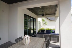 Modern patio area with a wooden dining table, grey chairs, and a seating area with blue cushions under a ceiling fan. Glass doors crafted by Texas builders open to the interior.