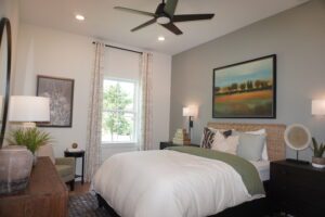 A modern bedroom featuring a neatly made bed, a ceiling fan, and decorative paintings on the walls, with natural light streaming in from a window. This space was crafted by Texas builders.