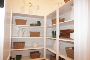 A well-organized pantry with white shelves holding wicker baskets, white and green dishes, and several pitchers, crafted by Texas builders.
