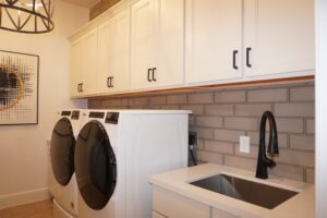 A modern laundry room with white cabinets, gray backsplash, a washing machine, dryer, and a utility sink designed by Texas builders.