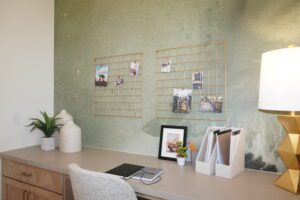 Modern home office designed by Texas builders with a clean desk, white chair, geometric lamp, minimalistic decor, and two photo grids on a textured green wall.