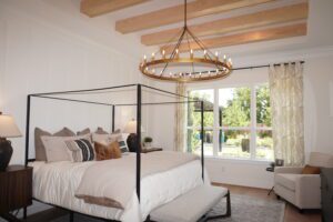 Modern bedroom designed by Texas builders, featuring a black four-poster bed, white linens, and wooden ceiling beams, with a circular chandelier and large windows.