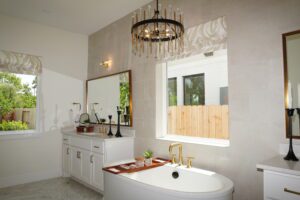 Modern bathroom interior featuring a freestanding bathtub, double vanity, and a circular chandelier, constructed by Texas builders, with a view of greenery through the window.