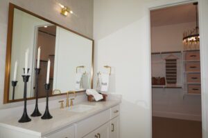 Modern bathroom with marble countertop, gold fixtures, and a mirror, crafted by Texas builders, leading to a walk-in closet with wood shelves and a brass chandelier.
