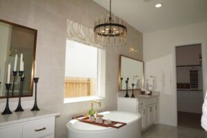 A modern bathroom designed by Texas builders, featuring a white bathtub, dual-sink vanity, and a unique chandelier, with decorative candles and mirrors.