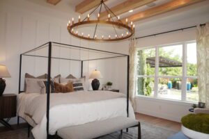 Elegant bedroom featuring a four-poster bed with white bedding, wooden side tables, a circular chandelier, and a view of the garden through large windows designed by Texas builders.
