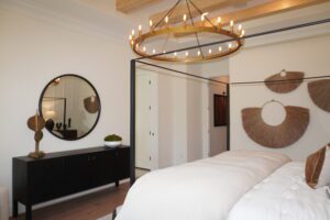 Modern bedroom featuring a large bed with white bedding, a black sideboard, a round mirror, circular light fixture, and decorative wall hangings crafted by Texas builders.