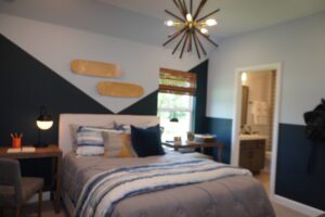 Modern bedroom designed by Texas builders featuring a geometric blue and gray wall, a bed with blue bedding, skateboard wall decor, and a sputnik chandelier. A bathroom is visible in the background.