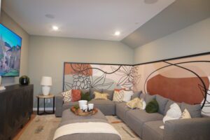 A cozy living room featuring a large gray sectional sofa adorned with various cushions, a mural wall with abstract floral designs, and a colorful painting by Texas builders.