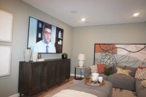 Modern living room with a large TV screen showing a man, gray sofa, wooden cabinet by Texas builders, and a decorative floral mural on the wall.