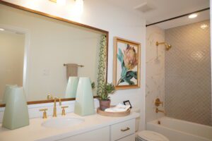 A clean, well-lit bathroom featuring a white vanity with dual sinks, gold fixtures, a large mirror, decorative vases, and a framed floral painting next to a glass-enclosed shower designed
