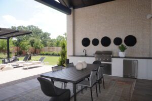 Modern outdoor patio featuring a dining area with a table and chairs, a built-in grill by Texas builders, and a lounge area with sunbeds, surrounded by a well-manicured lawn.